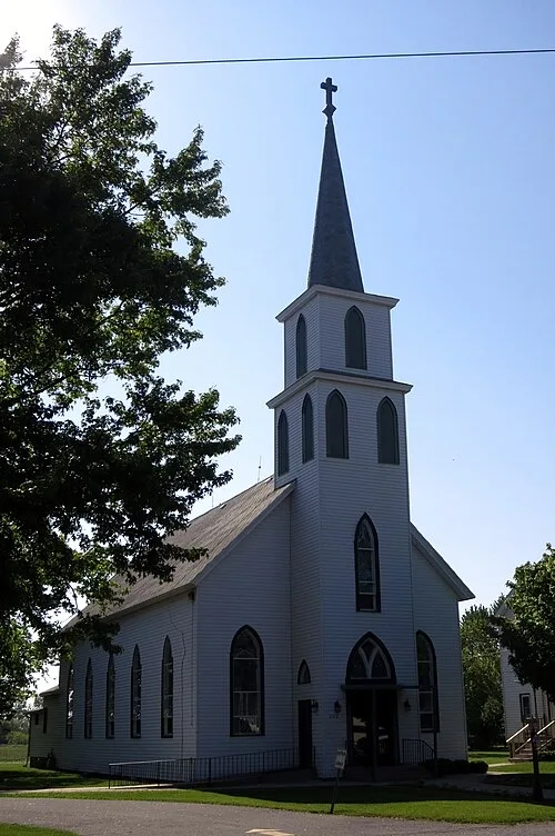 A white clapboard church with a tall steeple in Wanatah, Indiana, set against a clear summer sky.
